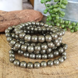May include: Several strands of pyrite gemstone bead bracelets. The beads are a metallic gray color and are strung together. The bracelets are displayed on a wooden surface, with a piece of wood and greenery in the background.