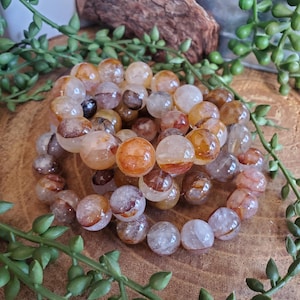 May include: A collection of round, translucent beads in shades of orange, brown, and clear. The beads are arranged in a circular pattern, suggesting a bracelet or necklace. The beads have a polished, glossy appearance, and are set against a wooden surface.