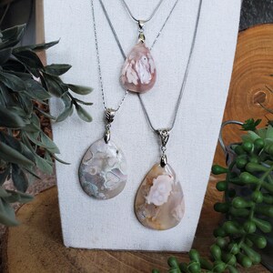 May include: Three gemstone pendant necklaces displayed on a white stand. The pendants are teardrop-shaped, featuring a mix of pink, white, and beige tones. Each necklace has a silver-colored chain. The background includes greenery and a wooden surface.