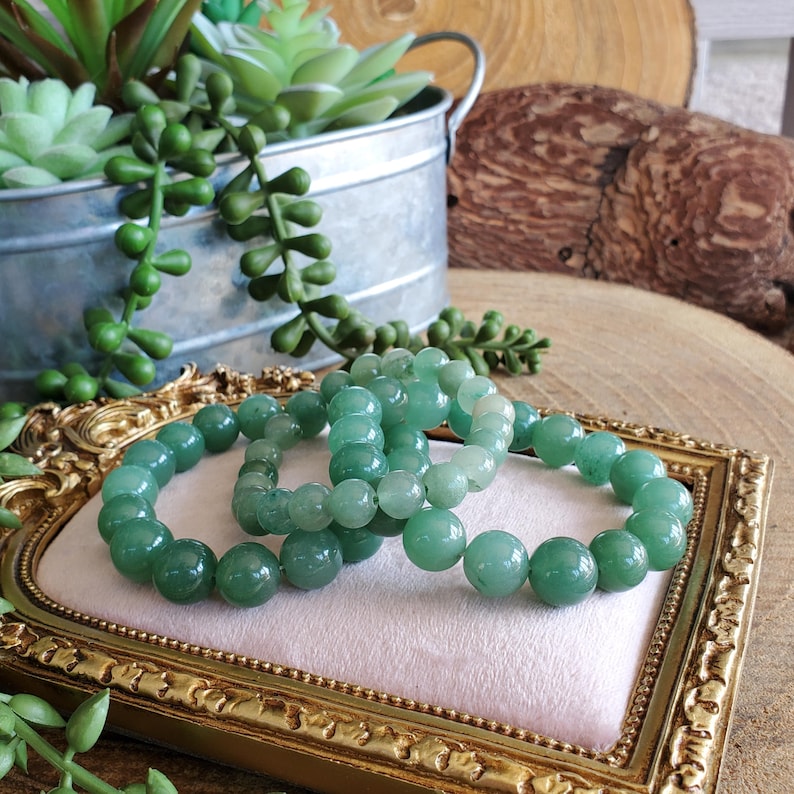May include: Three jade green beaded bracelets rest on a pink surface within a gold-framed display. The bracelets are composed of smooth, round beads. The background includes a metal container with greenery and a wooden slice.