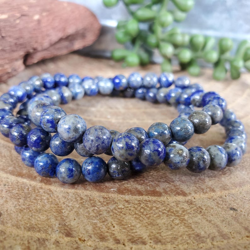 May include: Three strands of blue lapis lazuli gemstone beads. The beads are round and have a speckled appearance with shades of blue and gray. The strands are arranged in a circular pattern, showcasing the natural variations in the stone.
