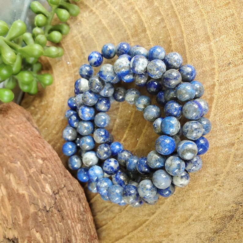 May include: A collection of blue and gray beaded bracelets. The beads are round and have a speckled appearance. The bracelets are arranged in a circular pattern on a wooden surface, with green foliage in the background.