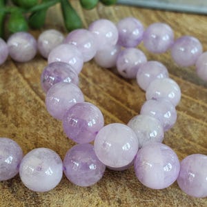 K&ouml;nnte beinhalten: Eine Kette aus runden, hellvioletten Amethystperlen. Die Perlen haben eine polierte, glatte Oberfl&auml;che und variieren in der Farbe von blassem Lavendel bis zu tieferem Violett. Die Perlen sind aufgef&auml;delt, was auf ein Armband oder eine Halskette hindeutet.