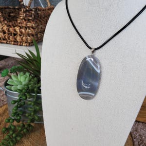 May include: A silver pendant necklace with a large oval-shaped agate stone. The stone has a white and gray banded pattern. The necklace is on a black cord.