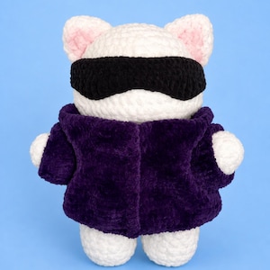 May include: A handmade crochet plush toy of a white cat-like creature wearing a black blindfold and a purple jacket. The ears are white with pink inner details. The toy is standing on a light blue background.