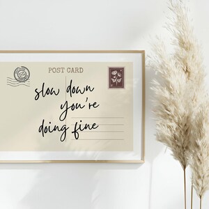 May include: A framed postcard print with the words "slow down you're doing fine" in black script. The postcard has a vintage aesthetic with a stamp and postmark design. The frame is a light gold color.