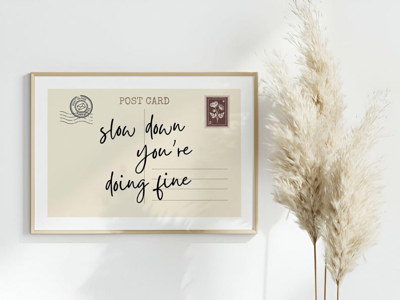 May include: A framed postcard print with the words "slow down you're doing fine" in black script. The postcard has a vintage aesthetic with a stamp and postmark design. The frame is a light gold color.