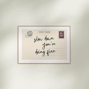 May include: A framed postcard print with the words "slow down you're doing fine" in black script. The postcard has a vintage aesthetic with a postage stamp and postmark design.