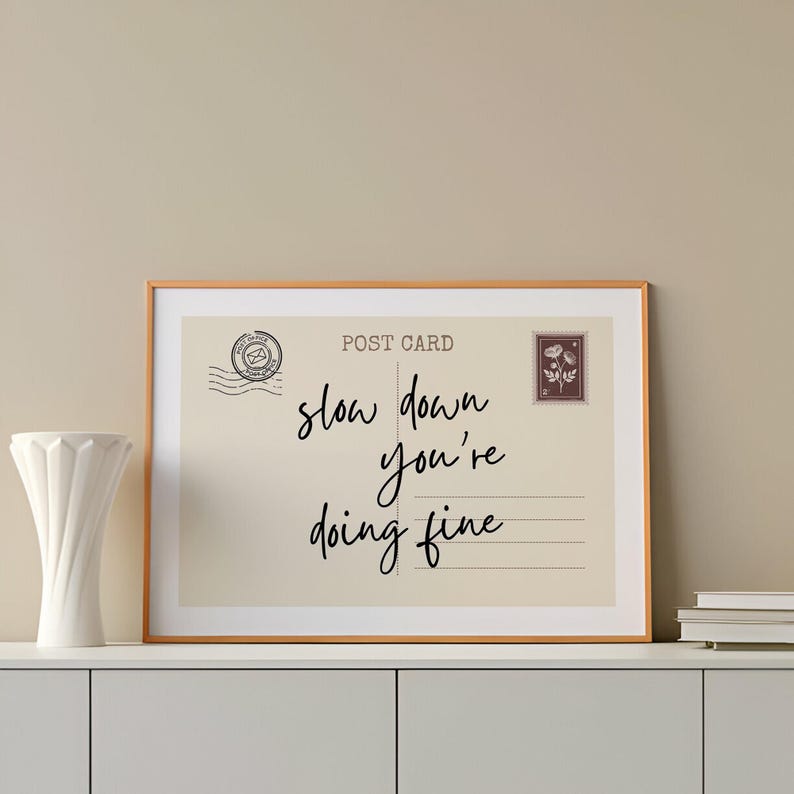 May include: Framed postcard print with the message "slow down you're doing fine" in black script. The print has a vintage aesthetic, with a postage stamp and postmark design. The frame is a warm, light brown color.