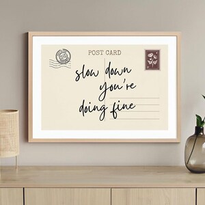 May include: A framed print with the words "slow down you're doing fine" in a handwritten style. The print resembles a postcard with a stamp and is displayed on a wooden surface with a lamp and vase.
