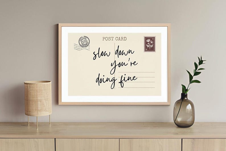 May include: A framed print with the words "slow down you're doing fine" in a handwritten style. The print resembles a postcard with a stamp and is displayed on a wooden surface with a lamp and vase.