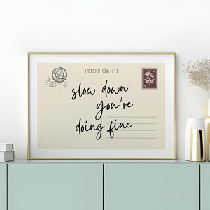 May include: A framed print resembling a vintage postcard, featuring the words "slow down you're doing fine" in elegant script. The print has a beige background with a gold frame, and includes a postage stamp and postmark.