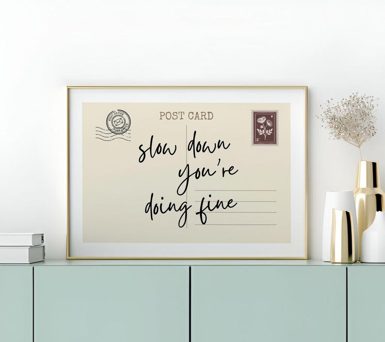 May include: A framed print resembling a vintage postcard, featuring the words "slow down you're doing fine" in elegant script. The print has a beige background with a gold frame, and includes a postage stamp and postmark.