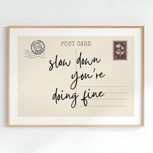 May include: A framed print featuring a vintage postcard design with the words "slow down you're doing fine" in elegant script. The postcard has a postage stamp and postmark details, all within a light beige background.