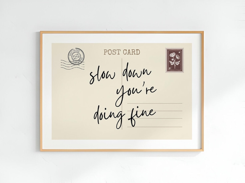 May include: A framed print featuring a vintage postcard design with the words "slow down you're doing fine" in elegant script. The postcard has a postage stamp and postmark details, all within a light beige background.