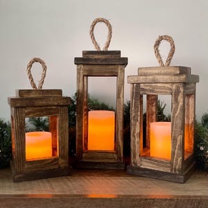 May include: Three wooden lanterns with rope handles. Each lantern has a rectangular frame with a warm, glowing candle inside. The wood has a dark, rustic finish, and the lanterns are arranged on a wooden surface.