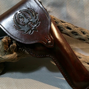 May include: A brown leather holster with a tooled design of a wolf's head. The holster is resting on a piece of driftwood.