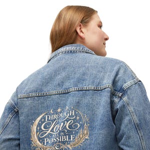 Puede incluir: Una chaqueta de mezclilla azul claro con un gráfico en la espalda que muestra una luna creciente, estrellas y la silueta de un castillo. El texto "THROUGH LOVE ALL IS POSSIBLE" está escrito en una fuente decorativa con detalles dorados y blancos.