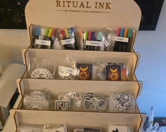 May include: A light-coloured wooden four-tiered display stand with the text RITUAL INK BY GODDESS STUDIO DESIGNS at the top. The shelves hold various small plastic-wrapped items, including colourful markers labelled Misc, Beltane, and Zodiac, and stickers.