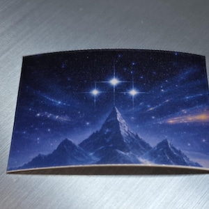 Puede incluir: Un posavasos rectangular con un paisaje celestial. El diseño muestra una cordillera bajo un cielo estrellado, con tres estrellas brillantes en un lugar destacado. La paleta de colores incluye tonos de azul, blanco y negro.