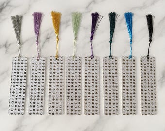 May include: Eight rectangular metal bookmarks arranged in a row, each featuring a grid of circular cutouts. Each bookmark is topped with a different coloured silk tassel: silver, purple, gold, light green, dark purple, dark green, blue, and black.