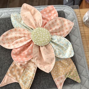 May include: A fabric flower ornament featuring a central circular piece in light green with a white floral pattern, surrounded by layered petals in various shades of peach, pink, and light blue with small floral and geometric prints, and two bottom tails.