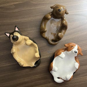 May include: Three ceramic dog-shaped dishes in brown, tan, and black. Each dish is designed to look like a dog lying on its back, with a shallow bowl for holding small items. The dogs have smiling faces.