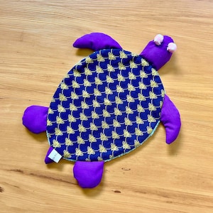May include: A plush turtle toy featuring a dark blue shell patterned with small yellow bees. The head, four flippers, and tail are made of solid purple fabric. The turtle has two small white circular eyes on its head and a white fabric tag.