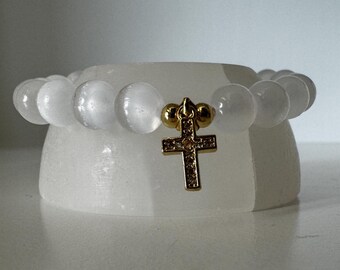 Op de afbeelding: Witte edelsteen armband met een gouden kruis bedel. Het kruis is bezet met kleine, heldere stenen.