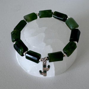 Könnte beinhalten: Ein Armband aus grünem Jade-Stein mit silbernen Perlen und einem silbernen Kaktus-Anhänger. Das Armband besteht aus rechteckigen Perlen.