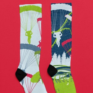 Könnte beinhalten: Ein Paar weiße und blaue Socken mit einem Fallschirmspringen-Motiv. Eine Socke zeigt eine grüne, blaue und weiße Fallschirmspringen-Szene. Die andere Socke zeigt eine grüne, blaue und weiße Waldszene.