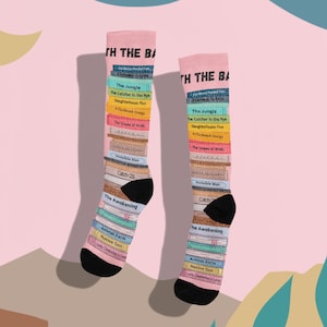 May include: A pair of pink socks with a design of stacked books featuring the titles of famous novels. The socks have black toes and heels. The text on the socks reads "With the Books".
