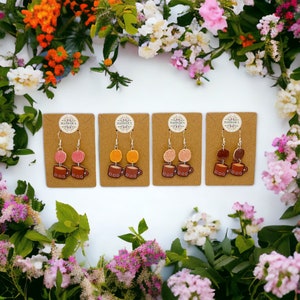 May include: Four pairs of dangle earrings featuring a brown coffee mug with a colorful swirl on top. The earrings are displayed on brown cards with the text "Handmade's" on each card.