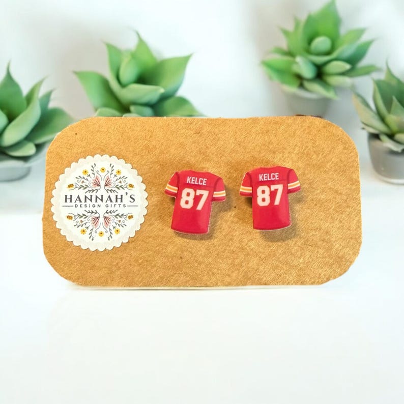 Op de afbeelding: Een paar rode en witte voetbaltrui-oorbellen met de naam "Kelce" en het nummer "87" erop. De oorbellen liggen op een bruine kaart met een klein logo waarop "Hannah's Design Gifts" staat.