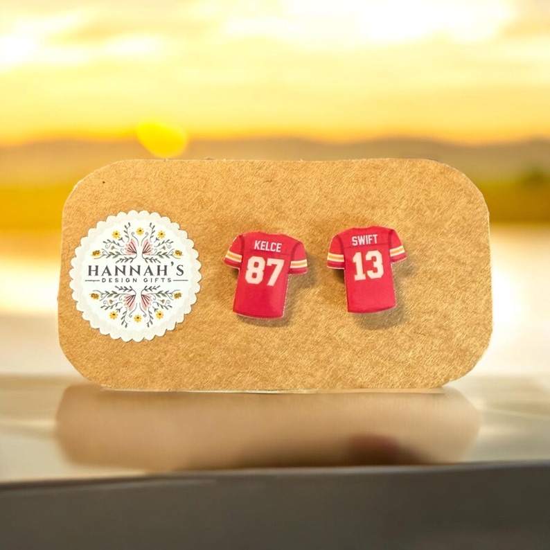 Op de afbeelding: Een paar rode en witte voetbaltrui-oorbellen met de nummers 87 en 13. De oorbellen zijn op een bruine kaart met de tekst "HANNAH'S DESIGN GIFTS" en de namen "KELCE" en "SWIFT" op de truien.