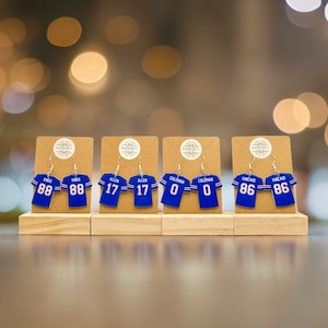 May include: Six pairs of blue and white football jersey earrings with player names and numbers on each jersey. The earrings are displayed on a wooden stand.