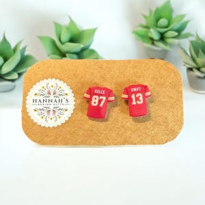 Op de afbeelding: Een paar rode en witte voetbaltrui-oorbellen met de namen "Kelce 87" en "Swift 13" erop. De oorbellen zijn op een bruine kaart met de tekst "Hannah's Design Gifts" erop.