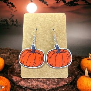 Op de afbeelding: Een paar oorhangers met oranje pompoenbedels en zilveren haakjes. De pompoenen hebben een steel en zijn zwart omrand.