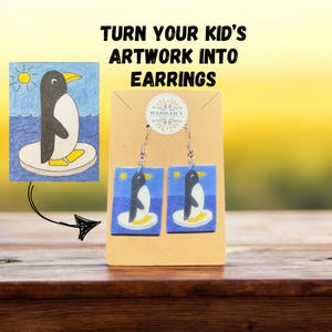 Op de afbeelding: Oorbellen met een pinguïnontwerp op een blauwe achtergrond. Ze worden gepresenteerd op een bruine kaart met de tekst "TURN YOUR KID'S ARTWORK INTO EARRINGS".