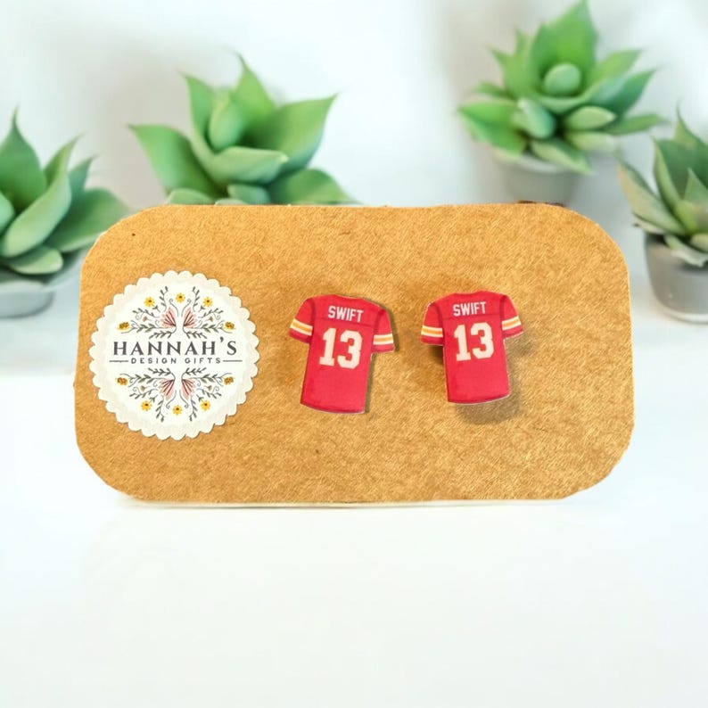 Op de afbeelding: Een paar rode en witte voetbaltrui-oorbellen met de naam "Swift" en het nummer "13" erop. De oorbellen worden getoond op een bruine kaart met een klein logo dat "Hannah's Design Gifts" zegt.