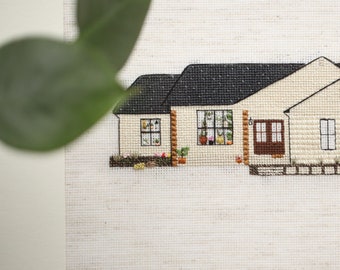 May include: A cross-stitch embroidery depicting a beige house with a black roof, brown door, and green bushes. The house features windows with white frames and a white picket fence.