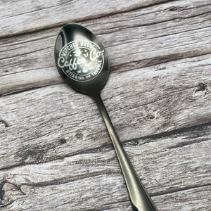 Może przedstawiać: Czarna metalowa łyżka z białym wzorem czarodziejskiego kapelusza i tekstem "Witches Brew Coffee Co. Est. 2023 Stirring Up Trouble"