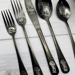 May include: A set of black stainless steel cutlery featuring the Grinch. The set includes a knife, fork, spoon, and a smaller spoon with the quote "Maybe Christmas doesn't come from a store. Maybe Christmas... perhaps... means a little bit more." The larger spoon features the Grinch and Max.