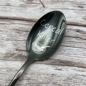 Könnte beinhalten: Ein schwarzer Löffel aus Metall mit den Worten "Cereal Killer" auf der Schüssel graviert. Der Löffel hat auch eine kleine Illustration einer Schüssel mit Müsli.