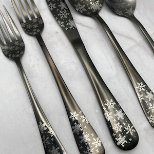 May include: A set of black silverware with snowflake designs. The set includes a fork, knife, and two spoons.