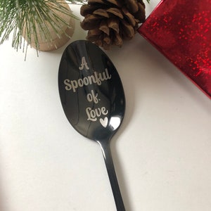 May include: A black spoon with the text "A Spoonful of Love" engraved on the spoon's head. A small heart is also engraved. The spoon is displayed with Christmas decorations, including pine cones and a red gift box.