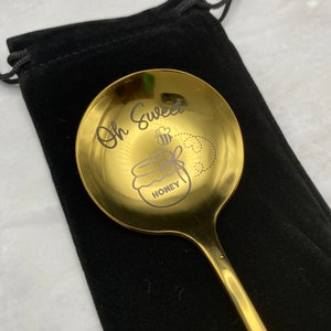 May include: A gold-colored spoon with a black velvet pouch. The spoon has the words "Oh Sweet" and a bee and honey jar design engraved on it.