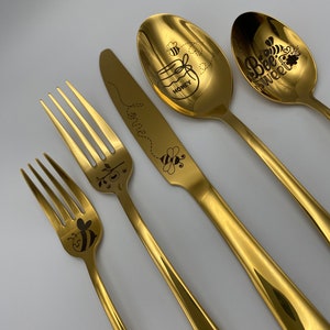 May include: A set of four gold-colored utensils with bee-themed designs. The utensils include a fork, a knife, and two spoons. One spoon has the text "Bee Sweet" and the other has the text "Honey" with a bee and a jar of honey.