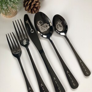 Puede incluir: Un juego de cinco cubiertos de metal negro con diseños grabados. Los cubiertos incluyen un cuchillo, dos tenedores y dos cucharas. Cada cubierto tiene la palabra "Alice" grabada en el mango.