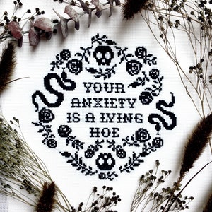 Może przedstawiać: Czarno-białe haftowane krzyżykiem z tekstem "Your anxiety is a lying hoe" otoczone wzorem kwiatowym z czaszkami i wężami.